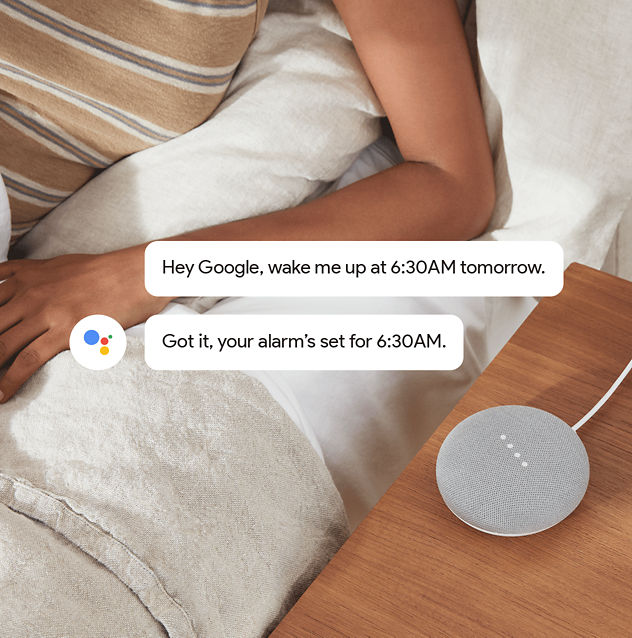 Google Home Mini con burbujas de comentarios arriba con una conversación. "OK Google, despiértame a las 6:30 a.m. mañana". "Comprendido, tu alarma está programada para las 6:30 a.m."